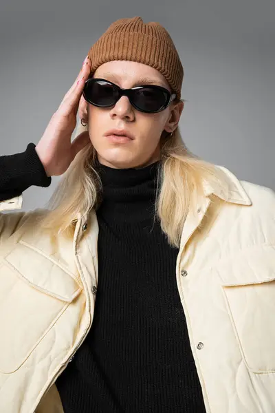Plano vertical de joven modelo no binario en gafas de sol y chaqueta de moda de invierno mirando a la cámara - foto de stock