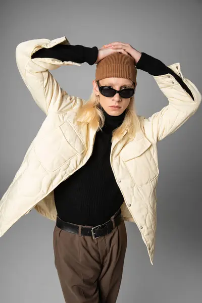 Joven persona no binaria en elegante chaqueta de invierno posando con las manos en la cabeza y mirando a la cámara - foto de stock