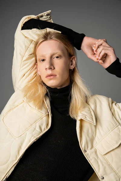 Attractive androgynous person in winter stylish jacket posing on gray backdrop with arms above head — Stock Photo