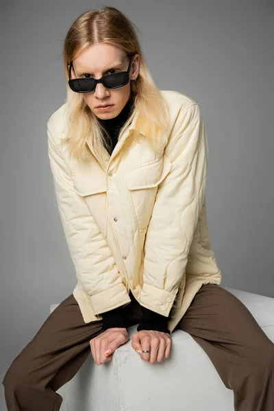 Scatto verticale di modello androgino alla moda in abito invernale seduto sul cubo e guardando la fotocamera — Foto stock