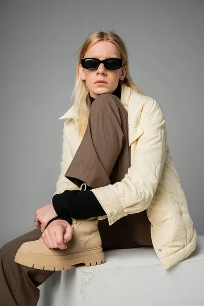 Gut aussehende androgyne Person in trendiger Winterkleidung auf riesigem White Cube sitzend, Modekonzept — Stockfoto