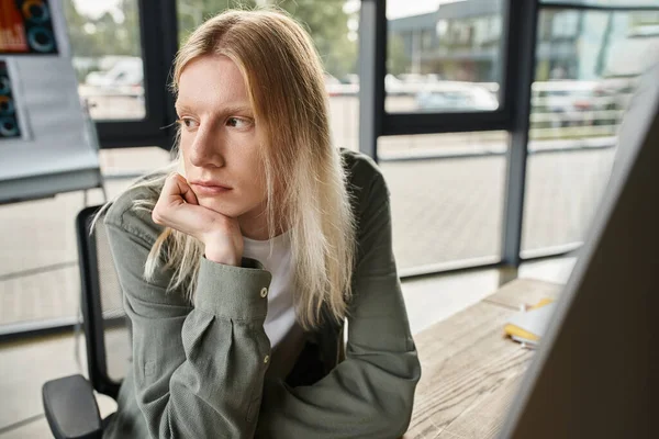 Pensive young androgynous person in green shirt sitting at desk with hand under chin, business — Stock Photo