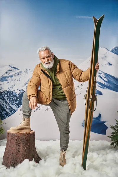 Fröhlicher Mann als Weihnachtsmann in sportlicher Kleidung legte sein Bein auf Baumstempel haltenden Ski, Weihnachtskonzept — Stockfoto