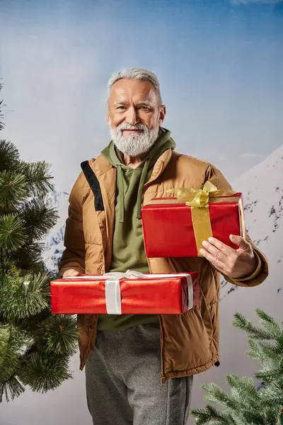 Cheerful man holding presents near pine tree with snowy mountains backdrop, winter concept — Stock Photo