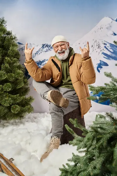 Feliz Papai Noel com barba branca sentado no toco da árvore com gesto símbolo de rocha, conceito de inverno — Fotografia de Stock