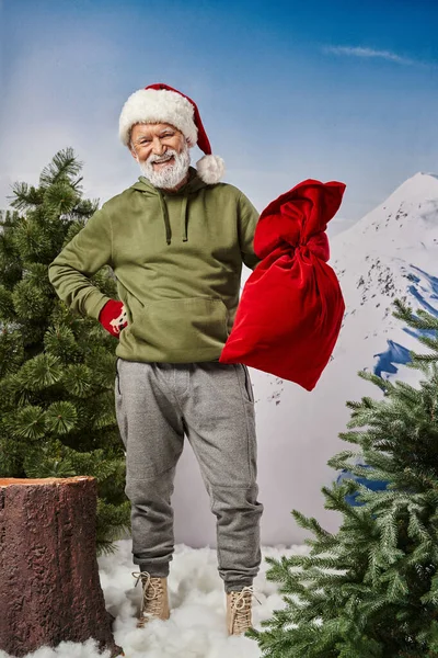 Homem alegre em cáqui com capuz segurando saco de Papai Noel e vestindo chapéu de Natal, Feliz Natal — Fotografia de Stock