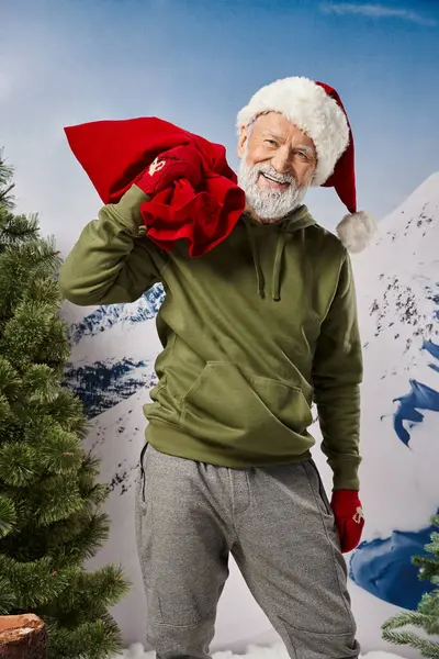 Santa alegre con sombrero navideño y bolsa de regalo sonriendo alegremente a la cámara, concepto de invierno - foto de stock