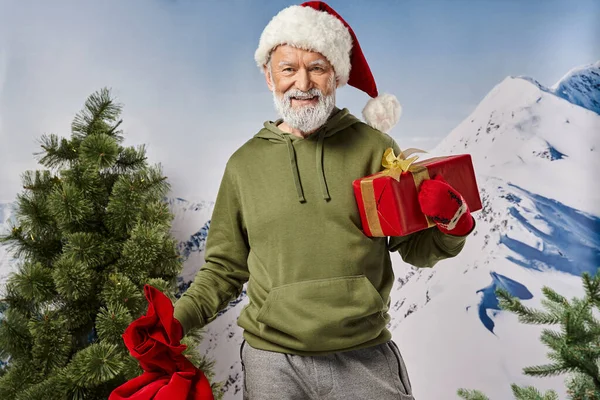 Alegre Santa en sombrero navideño con presente en la mano con fondo de montaña nevada, concepto de invierno - foto de stock