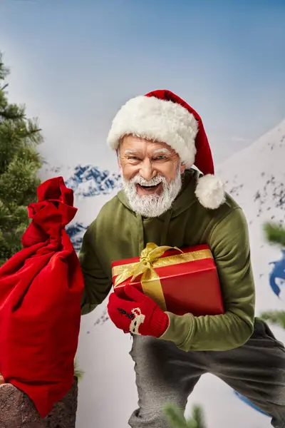 Alegre Santa con barba blanca en sombrero navideño con bolsa de regalo y regalo en la mano, concepto de invierno - foto de stock