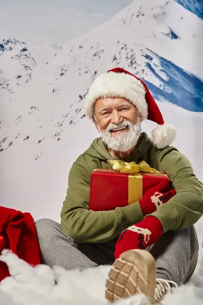 Joyful white bearded man dressed as Santa sitting on snow with present in hands, winter concept — Stock Photo