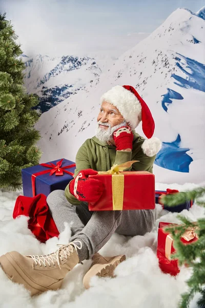 Мечтательный человек, одетый как Санта сидит на снегу в окружении подарков с рукой возле лица, зимняя концепция — стоковое фото