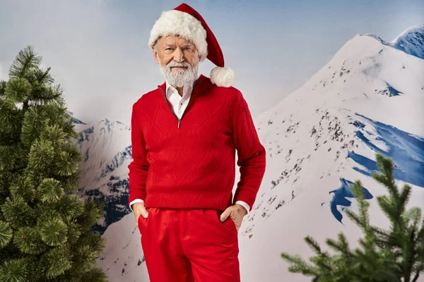Homem desportivo alegre em traje de Santa com as mãos em bolsos com fundo de montanha nevado, conceito de inverno — Fotografia de Stock