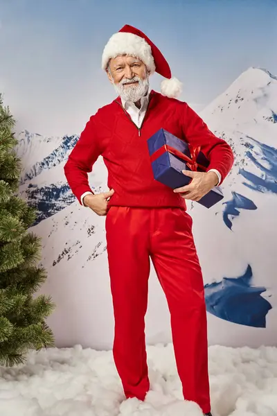 Fröhlicher Weihnachtsmann hält Geschenk mit einer Hand auf Hüfte vor verschneiter Bergkulisse, Winterkonzept — Stockfoto