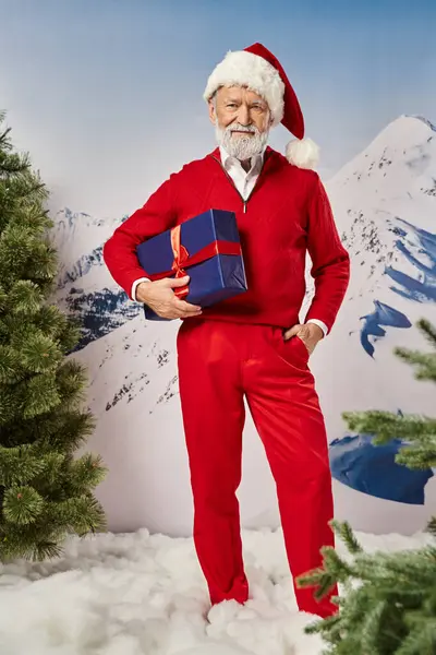 Стильний Санта позує з подарунком однією рукою в кишені з гірським тлом, зимова концепція — стокове фото