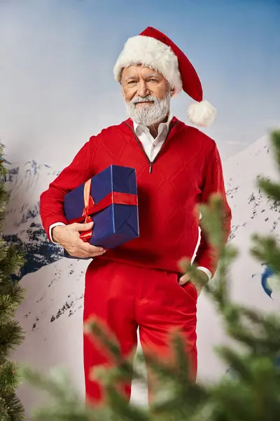 Homme élégant en costume de Père Noël avec cadeau et une main dans la poche près de sapin, concept d'hiver — Photo de stock