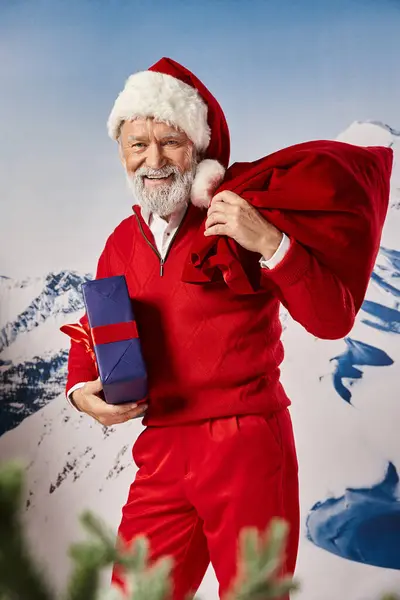 Jolly white bearded man in Santa costume posing with present bag with snowy backdrop, winter concept — Stock Photo