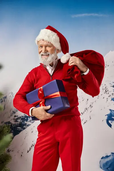 Hombre alegre en traje de Santa sonriendo sinceramente y sosteniendo regalo y bolsa de regalo, concepto de invierno - foto de stock