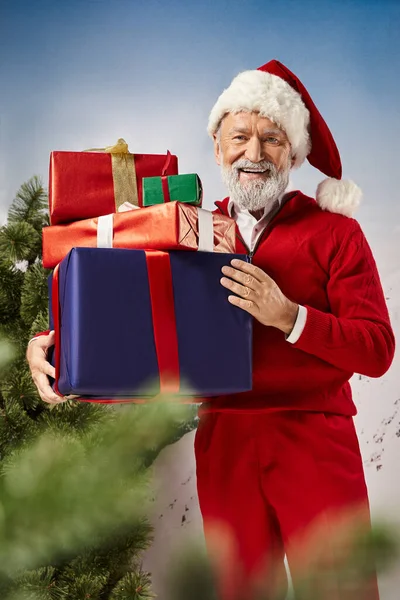 Hombre alegre en traje de Santa Claus rojo sosteniendo pila de regalos y sonriendo a la cámara, concepto de invierno - foto de stock
