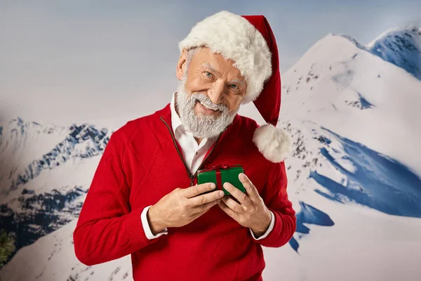 Alegre homem sorridente em Santa fantasia segurando pequeno presente com pano de fundo de montanha, conceito de inverno — Fotografia de Stock