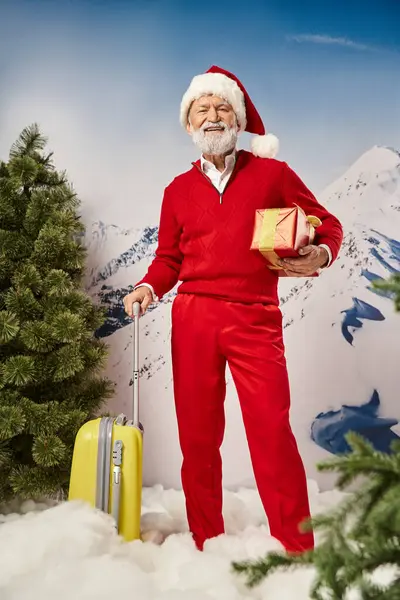 Happy man in Santa costume standing still with yellow suitcase and present in hand, winter concept — Stock Photo