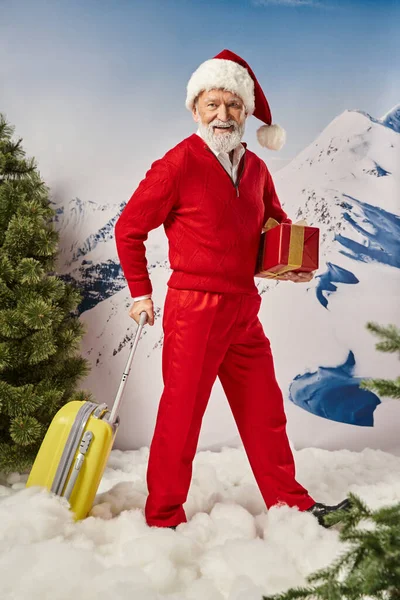 Stylish Santa with white beard holding present in hand posing with yellow suitcase, winter concept — Stock Photo