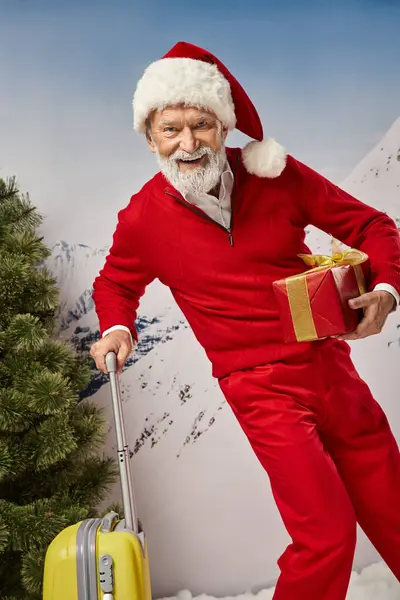 Веселый костюм Санты с белой бородой, позируя с желтым чемоданом и подарком, рождественская концепция — стоковое фото