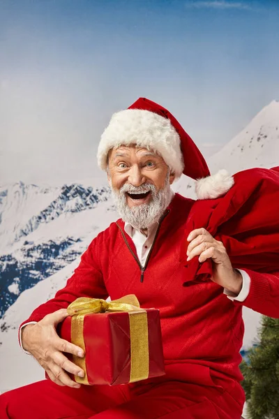 Alegre Santa en sombrero navideño con bolsa de regalo y presente sonriendo a la cámara, concepto de invierno - foto de stock