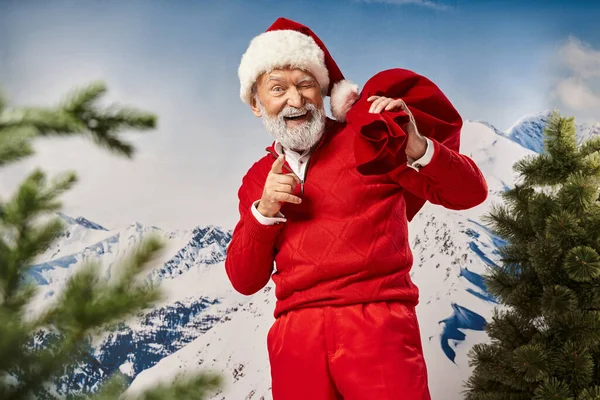 Playful bearded Santa with present bag winking and pointing finger at camera, Merry Christmas — Stock Photo