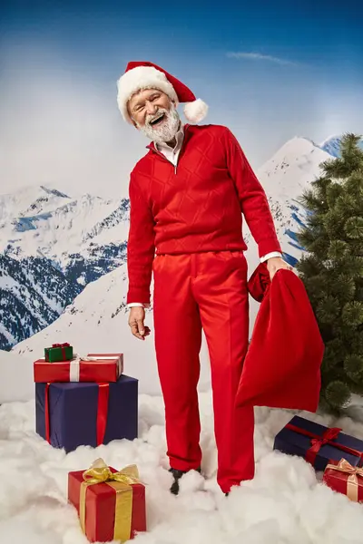 Cheerful man with gift bag surrounded by presents with snowy mountain backdrop, winter concept — Stock Photo