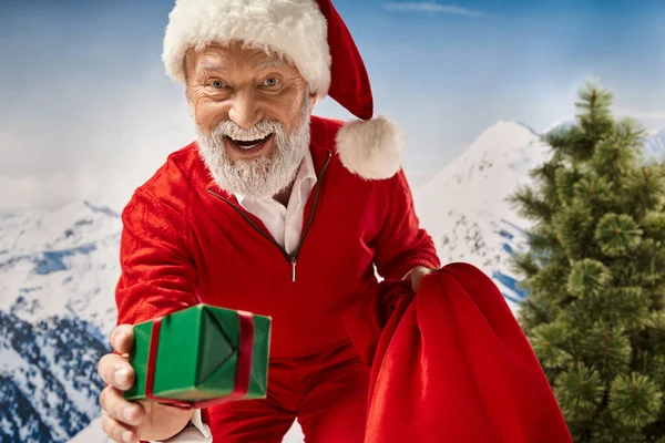 Homem alegre vestido de Santa emprestando presente na câmera e sorrindo alegremente, conceito de inverno — Fotografia de Stock