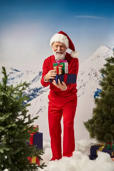Fröhliche Weihnachtsmänner mit Geschenken in den Händen, die vor verschneiter Bergkulisse in die Kamera lächeln, winterliches Konzept — Stockfoto