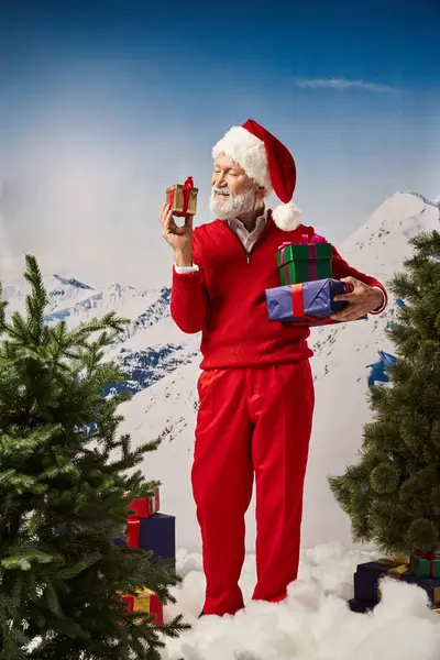 Радостный Санта в окружении подарков на снегу глядя весело на небольшой подарок, рождественская концепция — стоковое фото