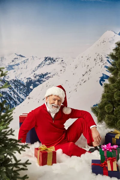 Jolly man dressed as Santa lying on snow surrounded by presents with snowy backdrop, winter concept — Stock Photo