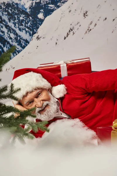 Close up of joyous Santa in christmassy hat lying on snow surrounded by presents, winter concept — Stock Photo