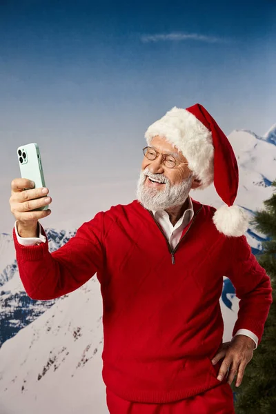 Cheerful Santa with glasses taking selfie with one hand on hip with snowy backdrop, winter concept — Stock Photo