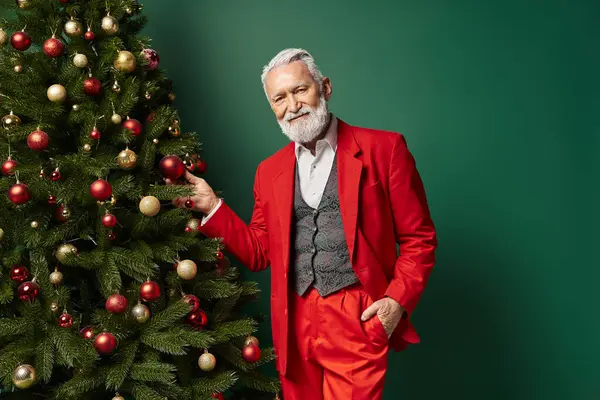 Eleganter, stilvoller Mann als Weihnachtsmann gekleidet, der neben dem Weihnachtsbaum vor grünem Hintergrund posiert, Winterkonzept — Stockfoto