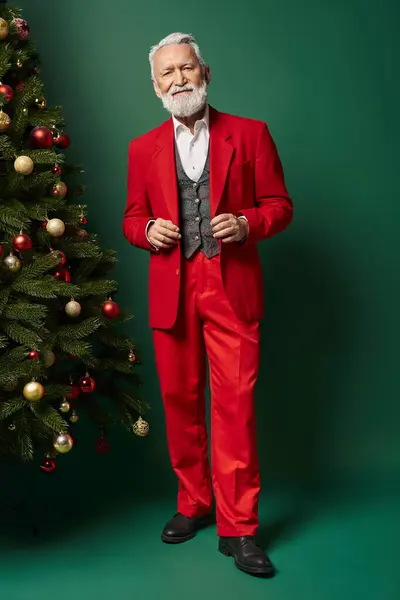 Guapo elegante Santa con barba blanca en traje rojo posando junto al abeto, concepto de invierno - foto de stock