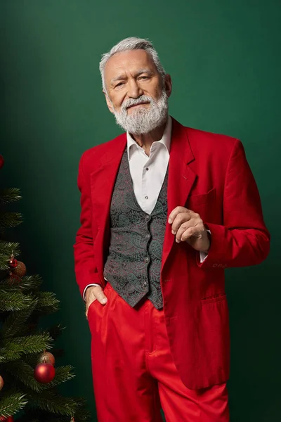 Elegante Santa con barba blanca con traje elegante y posando al lado de un pino, concepto de invierno - foto de stock