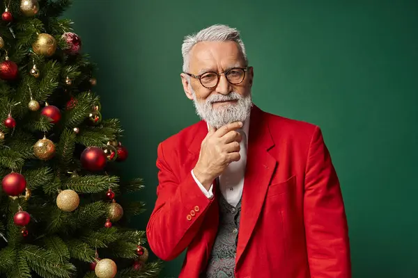 Fröhlicher Weihnachtsmann in Brille mit Bart im edlen Anzug posiert am Baum mit der Hand am Kinn, Winterkonzept — Stockfoto