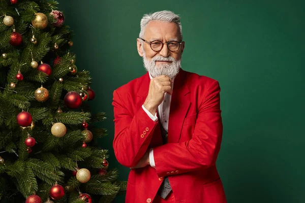 Щасливий Санта в стильному одязі з окулярами позує поруч з ялинкою з рукою на підборідді, зимова концепція — стокове фото