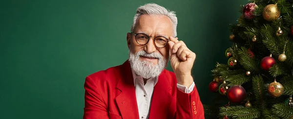 Élégant homme barbu dans des lunettes posant à côté de pin sur fond vert, concept de Noël, bannière — Photo de stock