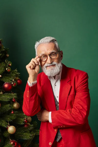 Alegre hombre con estilo en traje de Santa tocando gafas posando junto al árbol de Navidad, concepto de invierno - foto de stock