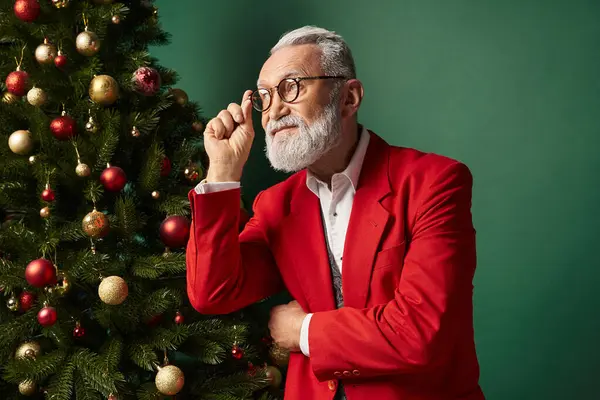 Bonito Santa em óculos de pé ao lado da árvore de Natal com bugigangas olhando para longe, conceito de inverno — Fotografia de Stock
