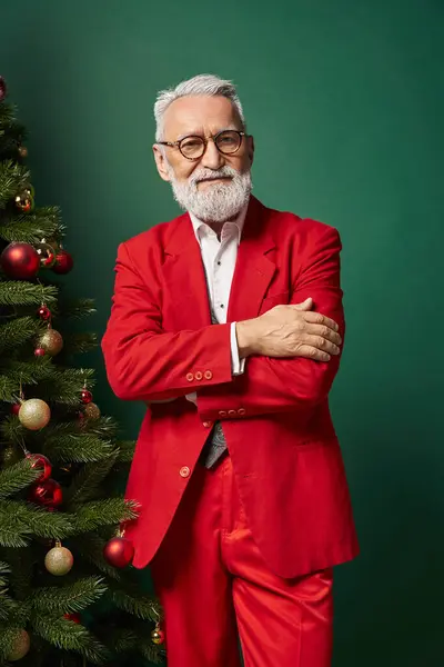 Élégant Père Noël en costume rouge avec des lunettes posant près de l'arbre de Noël avec les bras croisés, concept d'hiver — Photo de stock