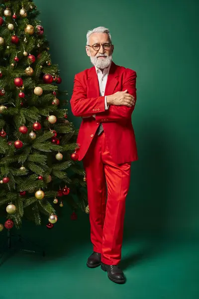 Schöner Weihnachtsmann mit weißem Bart und Brille, der neben Tanne mit verschränkten Armen posiert, winterliches Konzept — Stockfoto