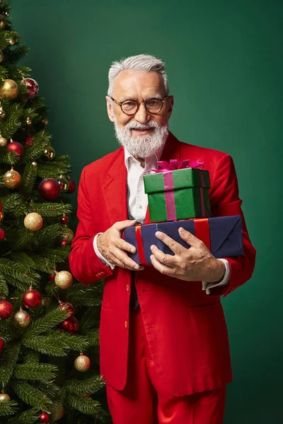 Elegante allegro Babbo Natale in abito rosso in posa accanto al pino che tiene mucchio di regali, concetto invernale — Foto stock