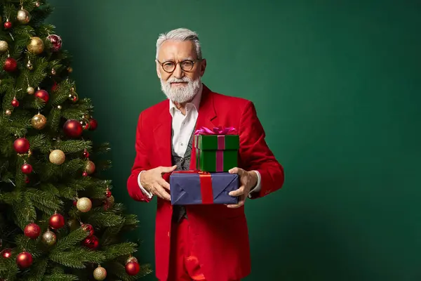Elegante Santa con gafas y barba sosteniendo pila de regalos sobre fondo verde oscuro, concepto de invierno - foto de stock
