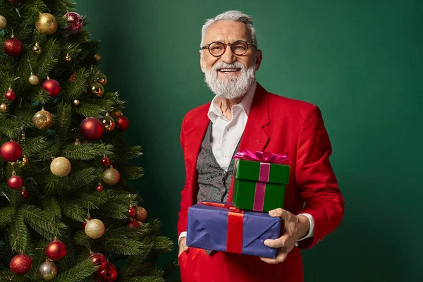 Веселий стильний чоловік, одягнений як Санта в окуляри, тримає подарунки поруч з ялинкою, зимова концепція — стокове фото