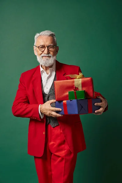 Красивий чоловік одягнений як елегантний Санта позує з подарунками на темно-зеленому тлі, зимова концепція — стокове фото