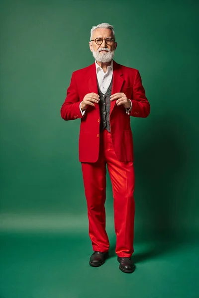 Serio elegante Santa posando con las manos en traje rojo sobre fondo verde oscuro, concepto de invierno - foto de stock